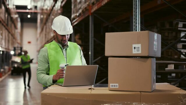 Male Worker Scanning Cardboard Box with Barcode Scanner and Using Laptop at Warehouse. Worker Placement Package on Shelf. Rows of Cardboard Boxes with Ready Products in Background