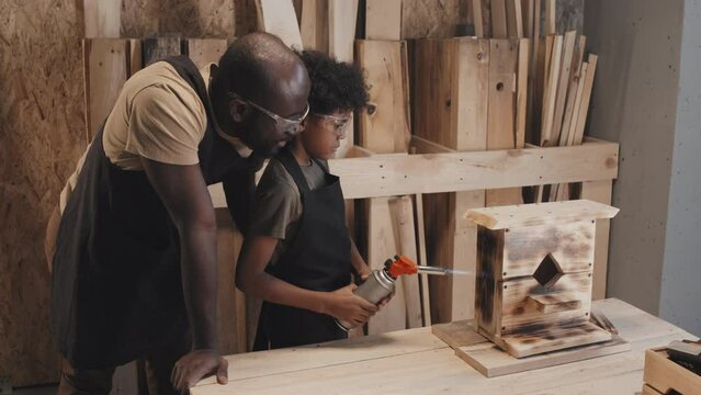 African American kid scorching handmade birdhouse together with father in carpentry studio