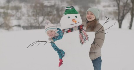 Young mother and small child has fun outside in winter and peek out from behind snowman and smile. Family in winter holidays