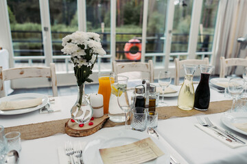 Table setting with wild flowers. Panoramic windows on the summer terrace overlooking the forest and the lake. Lifebuoys behind the pier. Restaurant on the water.