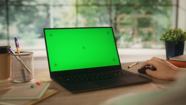 Over the Shoulder Footage with a Small Business Owner Working Remotely from Home, Using Laptop Computer with Mock Up Green Screen Display. Person Sitting Behind a Cozy Wooden Desk, Browsing Internet