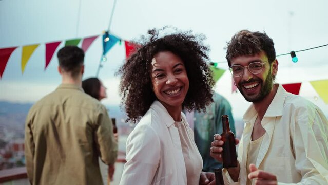 Portrait of young adult multiracial couple smiling, laughing and looking at camera holding beer bottles. Two people staring front on a party celebration with their best friends talking at background
