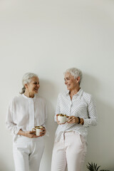 Senior females drinking coffee and laughing on white wall background.