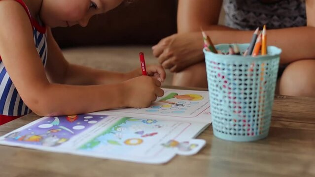 Cute little girl coloring pictures in a drawing book with her mother sitting next to her