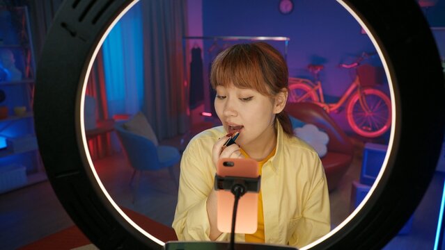 Attractive girl blogger in casual clothing applying a red lipstick and filming in front of the camera at home studio.