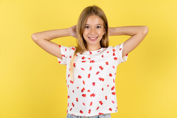 Satisfied blonde kid girl wearing polka dot shirt over yellow studio background hold hands behind...