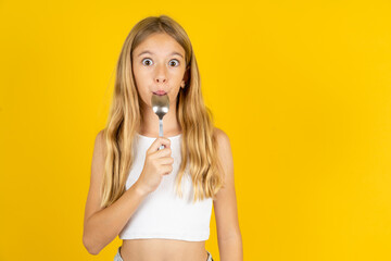 Very hungry blonde kid girl wearing white T-shirt over yellow studio background holding spoon into...