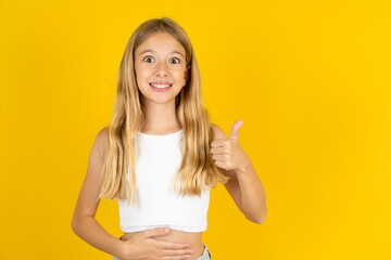 blonde kid girl wearing white T-shirt over yellow studio background happy positive smile hands on...