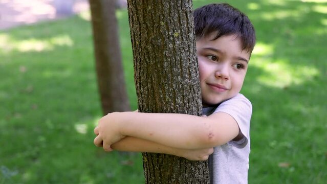 adorable cute smiling preschooler boy kid hugging a tree in park, green grass summertime. hug a tree day close up 4k