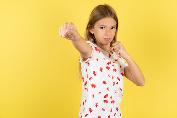 blonde kid girl wearing polka dot shirt over yellow studio background Punching fist to fight,...