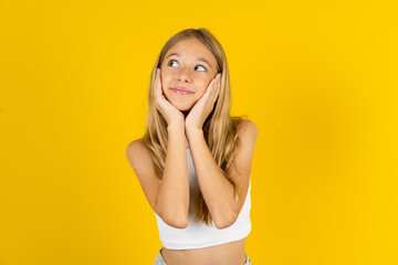 Inspired blonde kid girl wearing white T-shirt over yellow studio background looking at copyspace...