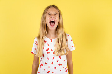 blonde kid girl wearing polka dot shirt over yellow studio background angry and mad screaming frustrated and furious, shouting with anger. Rage and aggressive concept.