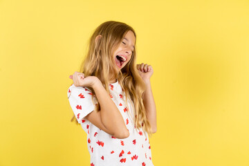 Ecstatic blonde kid girl wearing polka dot shirt over yellow studio background shout loud yeah fist...