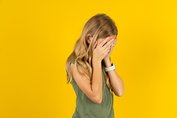 Sad blonde kid girl wearing green T-shirt over yellow studio background crying covering her face with her hands.
