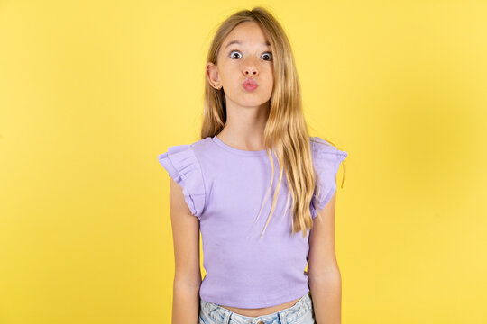 Shot of pleasant looking blonde kid girl wearing violet T-shirt over yellow studio background , pouts lips, looks at camera, Human facial expressions