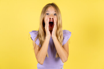 blonde kid girl wearing violet T-shirt over yellow studio background shouting excited to front.