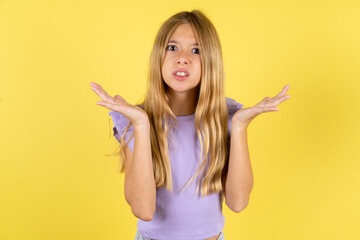 Indignant blonde kid girl wearing violet T-shirt over yellow studio background gestures in bewilderment, frowns face with dissatisfaction. - obrazy, fototapety, plakaty