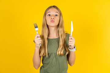 hungry blonde kid girl wearing green T-shirt over yellow studio background holding in hand fork...