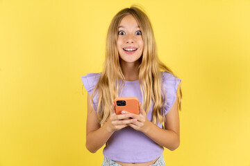 Excited blonde kid girl wearing violet T-shirt over yellow studio background holding smartphone and looking amazed to the camera after receiving good news.