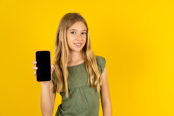 Smiling blonde kid girl wearing green T-shirt over yellow studio background Mock up copy space. Hold mobile phone with blank empty screen