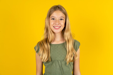 blonde kid girl wearing green T-shirt over yellow studio background with nice beaming smile pleased expression. Positive emotions concept