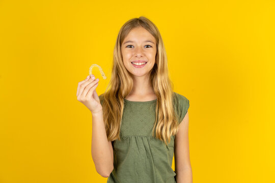 blonde kid girl wearing green T-shirt over yellow studio background holding an invisible braces aligner, recommending this new treatment. Dental healthcare concept.
