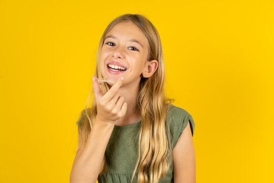 blonde kid girl wearing green T-shirt over yellow studio background holding an invisible aligner ready to use it. Dental healthcare and confidence concept.