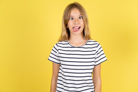 Funny young beautiful blonde kid girl over yellow studio background makes grimace and crosses eyes plays fool has fun alone sticks out tongue.