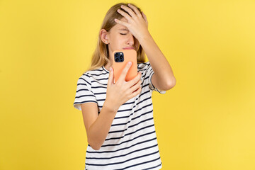 Upset depressed young beautiful blonde kid girl over yellow studio background makes face palm as forgot about something important holds mobile phone expresses sorrow and regret blames