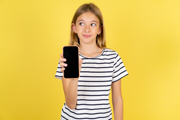 Fototapeta premium young beautiful blonde kid girl over yellow studio background holds new mobile phone and looks mysterious aside shows blank display of modern cellular