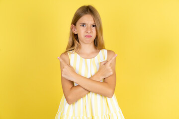 Serious young beautiful blonde kid girl over yellow studio background crosses hands and points at...
