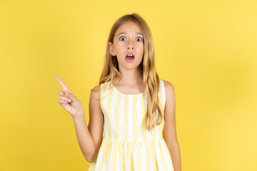 Emotive young beautiful blonde kid girl over yellow studio background keeps jaw dropped from shock...