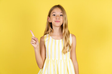 Positive young beautiful blonde kid girl over yellow studio background with satisfied expression indicates at upper right corner shows good offer suggests to click on link