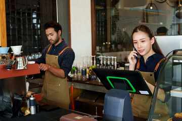 Husband and wife couple owning a small coffee shop business, Asian woman answering calls from regular customers and male barista preparing coffee behind bar counter where customers order.