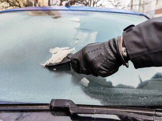 Cleaning car windows in winter. Man scrapes hoarfrost with a plastic scraper from the windshield of...