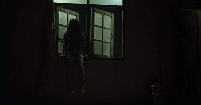 Horror scene of a mysterious Scary Asian ghost woman creepy have hair covering the face sitting on window at abandoned house with dark scene movie at night, festival Halloween concept