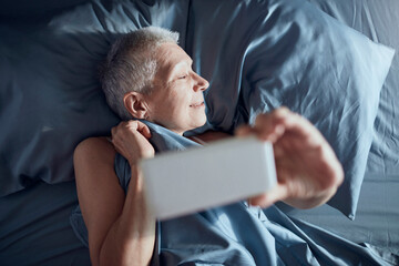 Senior woman taking a selfie with a smart phone in a bed in bedroom at home
