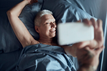 Senior woman taking a selfie with a smart phone in a bed in bedroom at home