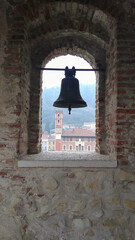 Obraz premium Bell in the tower window of an ancient castle. Construction of red bricks and stones..In the background, view of the city, buildings and green mountains.