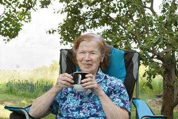 Portrait of happy smiling senior grandmother in garden at home. old woman over 80 rests sitting in chair in nature, drinking tea or water. Summer. Old age
