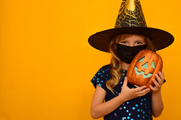 A little cute girl dressed as a Witch on an orange background in a black medical mask plays trick...