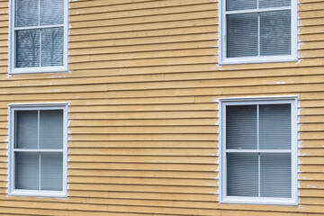 The exterior wall of a vintage yellow wooden house with four double hung window. The wall is pine cape cod siding with horizontal boards. The glass windows have white trim and white blinds hanging. 