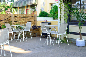 Fototapeta na wymiar Multiple empty white metal tables with round glass tops and white rattan chairs at a sidewalk cafe patio of a restaurant. The outdoor restaurant wall is made of brown lattice wooden boards. 