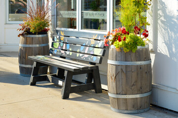 The exterior of a shop with a brown plastic bench painted in yellow, pink, and blue colors on a sidewalk. There are two wooden wine barrels next to the seating area with colorful flowers planted.  - Powered by Adobe