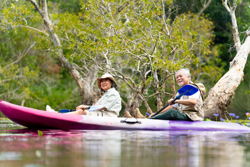 Asian senior couple kayaking together in the lake at mangrove forest on summer vacation. Retired elderly people man and woman have fun outdoor lifestyle travel nature and rowing a boat in the river.