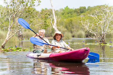 Asian senior couple kayaking together in the lake at mangrove forest on summer vacation. Retired elderly people man and woman have fun outdoor lifestyle travel nature and rowing a boat in the river.