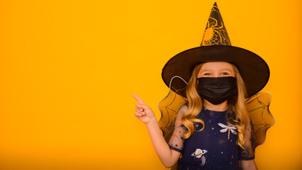 A little cute girl dressed as a Witch on an orange background in a black medical mask plays trick...