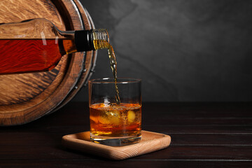 Pouring whiskey from bottle into glass with ice cubes near wooden barrel on table, space for text