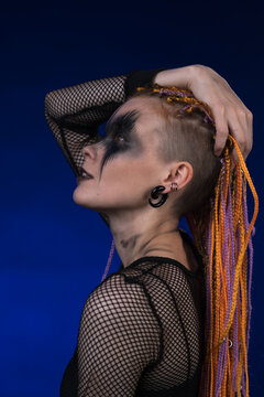 Cinematic portrait of informal woman with colored braids hairdo and horror black stage make-up painted on face. Studio shot on blue background. Part of photo series
