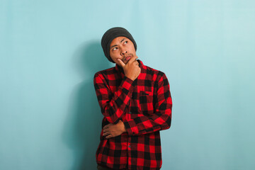 Thoughtful young Asian man with a beanie hat and a red plaid flannel shirt keeps his hand on his chin, looking aside at a copy space with a serious expression while standing against a blue background